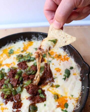 Hand holding a chip with queso fundido with chorizo and the melted cheese is dripping down into the appetizer dish.