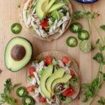 Two tostadas of Salpicon de Pollo topped with avocado slices and surrounded by cilantro leaves .and jalapenos.
