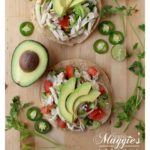 Two tostadas topped with Salpicon de Pollo topped with avocado slices surrounded by cilantro and jalapeno.