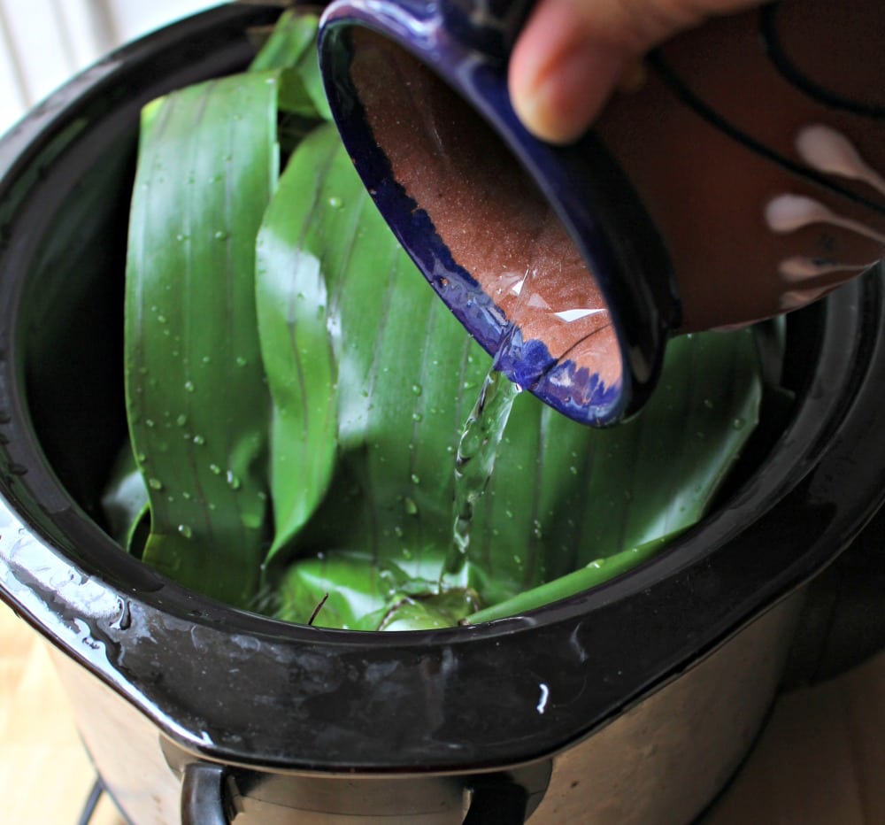 A clay cup pouring water into the slow cooker around the banana leaves.