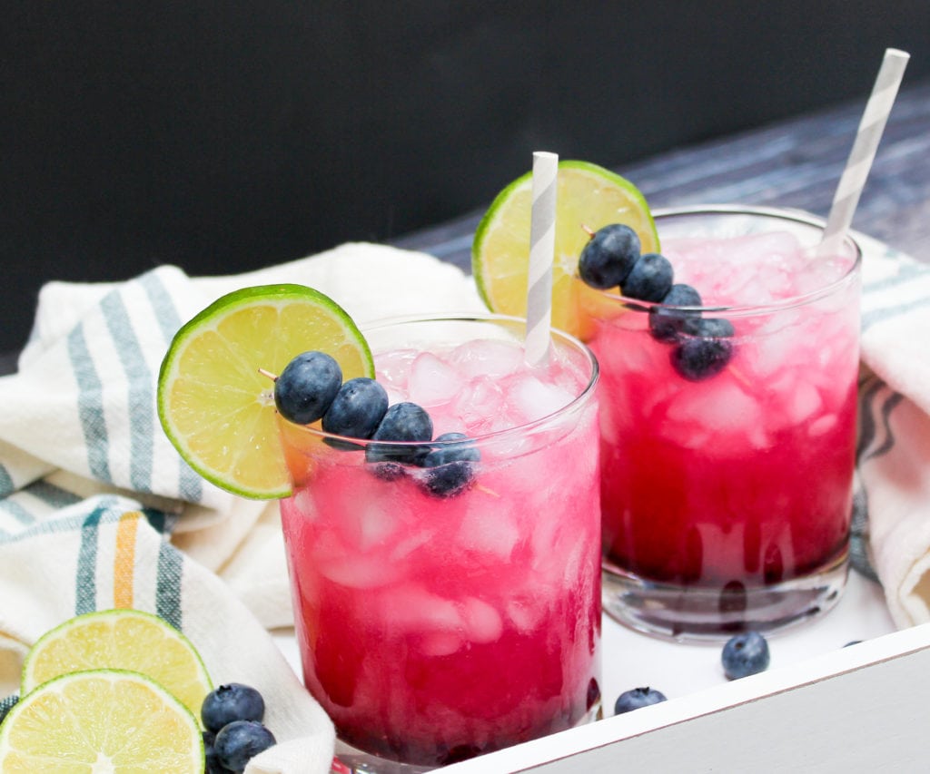 Two blueberry margaritas served with ice and topped with blueberries and lime wedges.