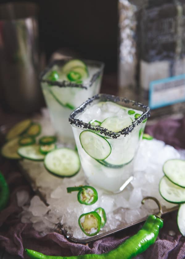 Serrano Cucumber Margarita served in a square glass topped with black salt, cucumber slices and serrano pepper slices.