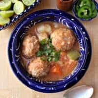 Albondigas en Caldillo (or Mexican Meatball Soup) in a decorative blue bowl next to lime and a spoon.