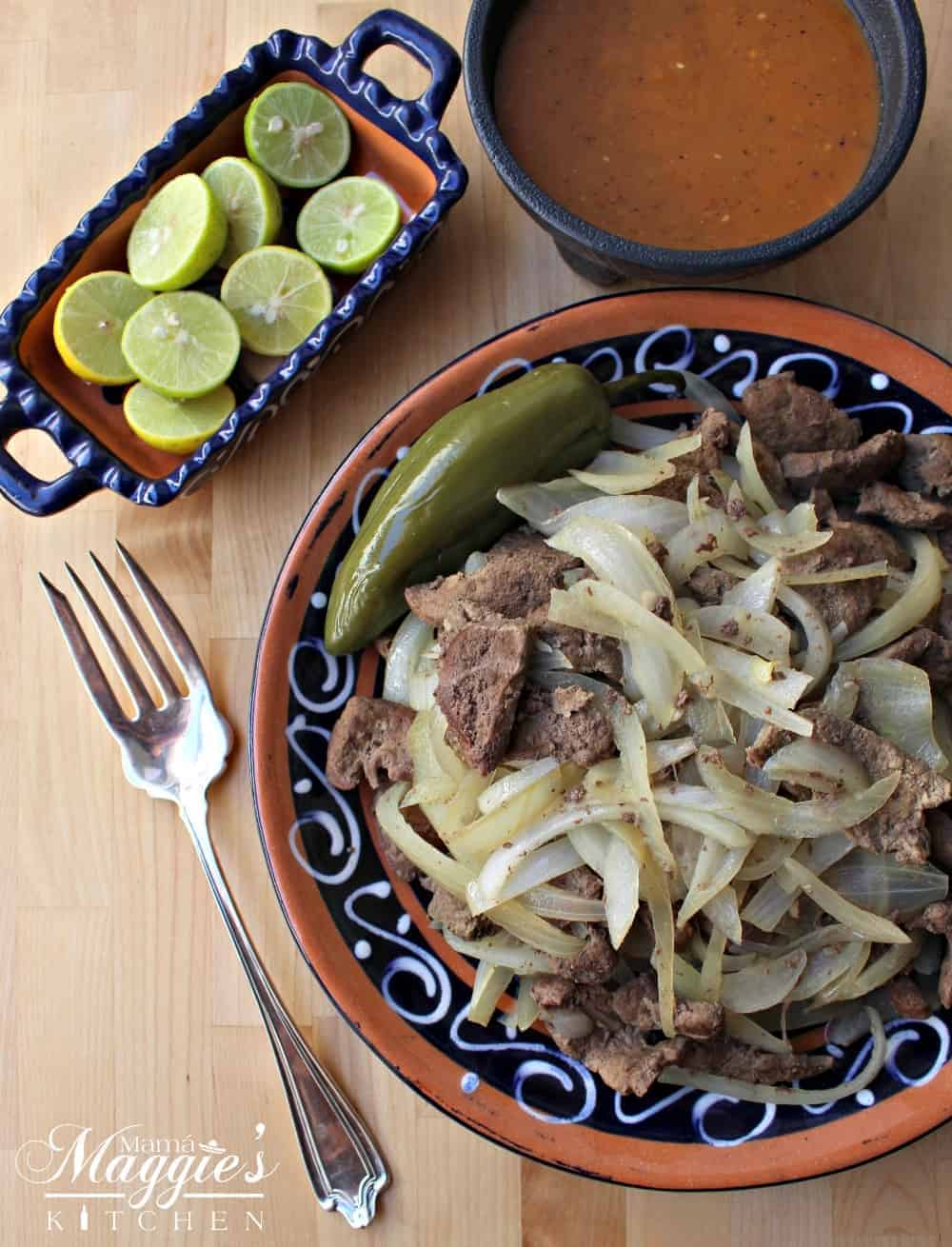 Beef liver and onions in a decorative Mexican plate next to a fork and limes.
