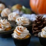 Pumpkin cupcakes topped with frosting and drizzled with cajeta next to a pine cone and an orange pumpkin.