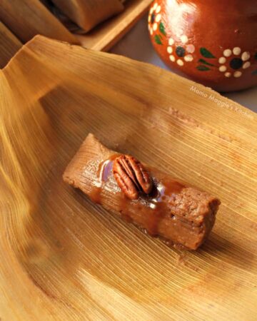 A tamal sitting on top of a corn husk drizzled with cajeta sauce and topped with a pecan next to a decorative clay mug.