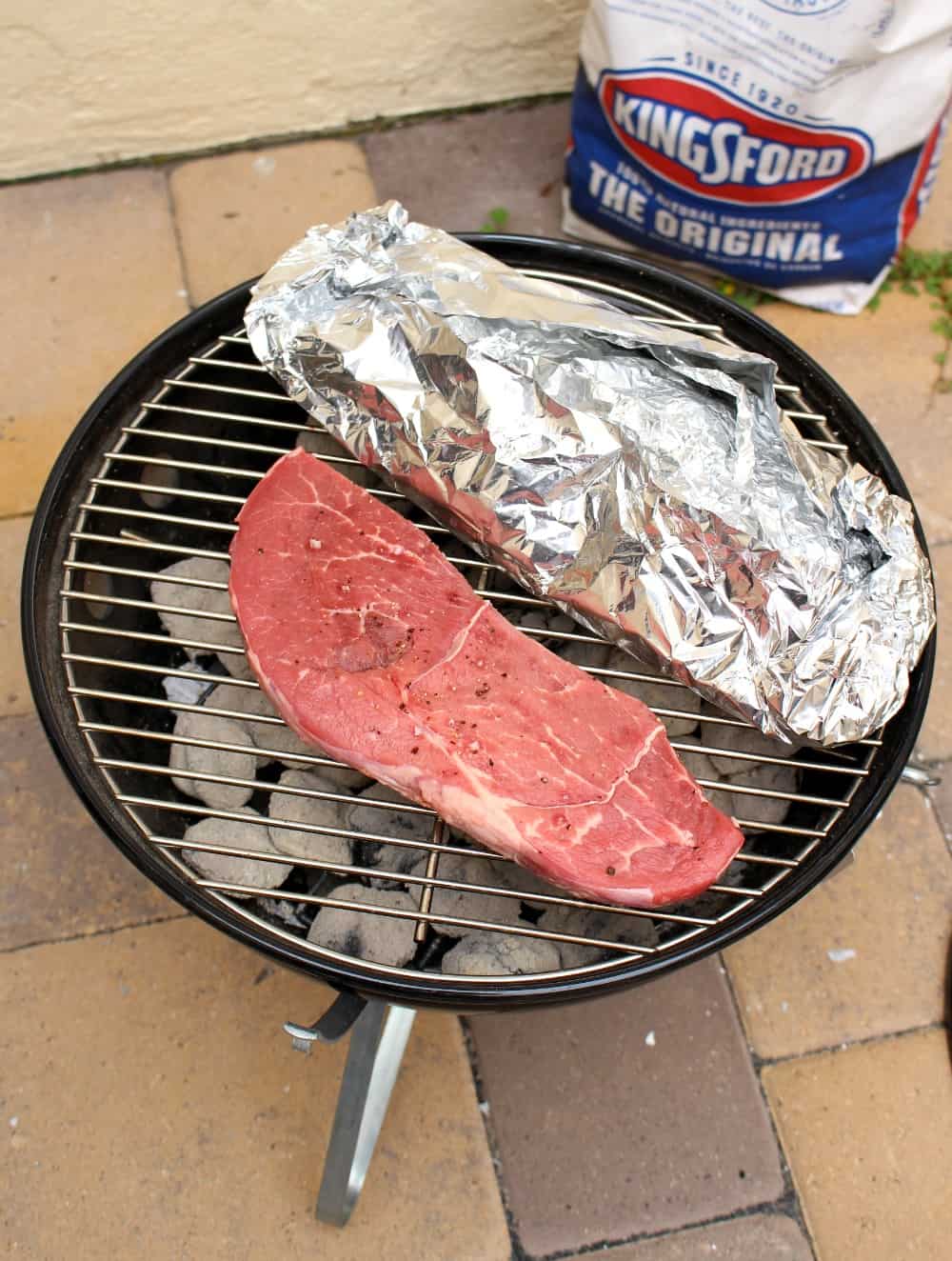 A steak cooking on a grill next to the potato foil packet. 