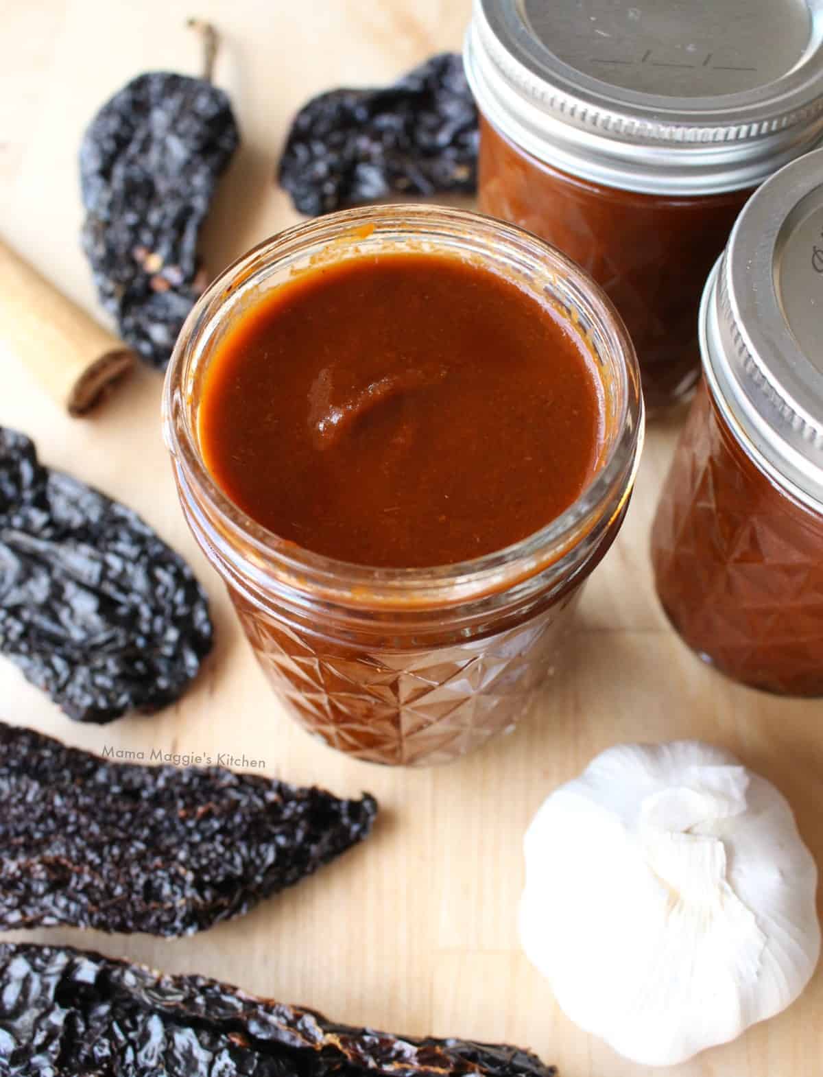 A jar of adobo sauce surrounded by ancho chiles and a bulb of garlic.