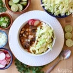 Mushroom vegan pozole rojo served in a white bowl toped with cabbage and radishes and surrounded by more toppings.