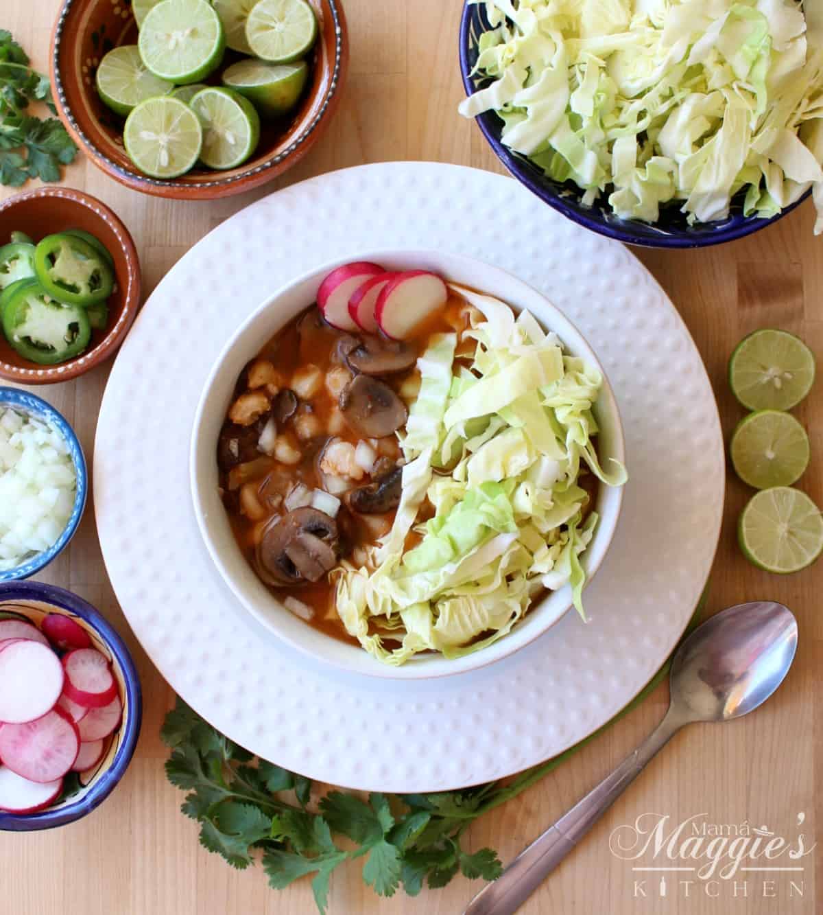 Mushroom vegan pozole rojo served in a white bowl toped with cabbage and radishes and surrounded by more toppings.
