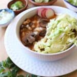 Vegan pozole rojo served in a white bowl topped with cabbage and sliced radishes surrounded by more toppings.