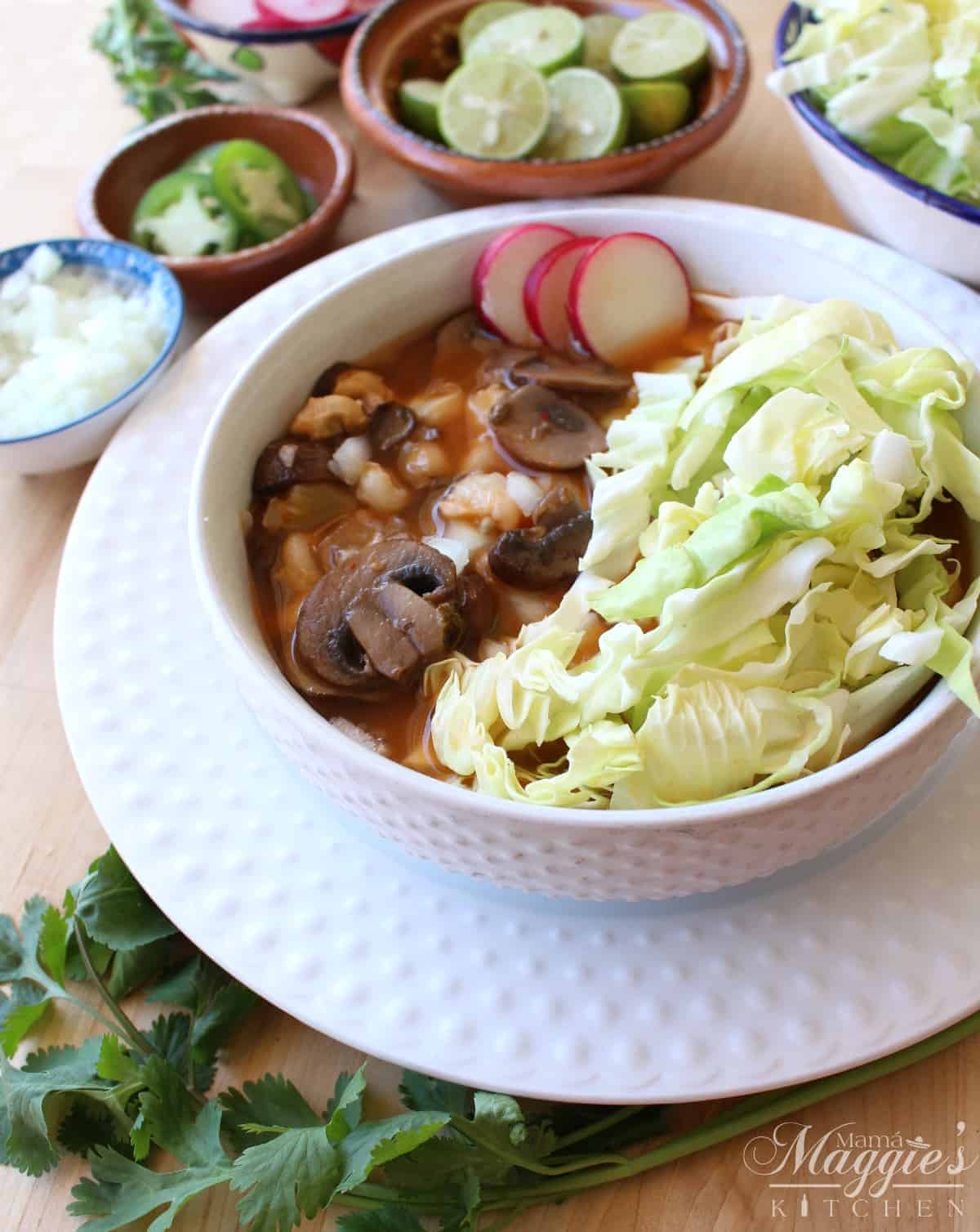 Vegan pozole in a white bowl topped with cabbage and sliced radishes surrounded by more toppings.