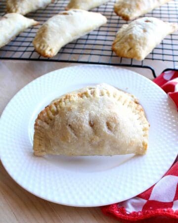 An apple empanada on a white plate next to a red checkered napkin and more empanadas cooling.