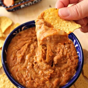 A hand holding a chip with frijoles puercos over a blue bowl.