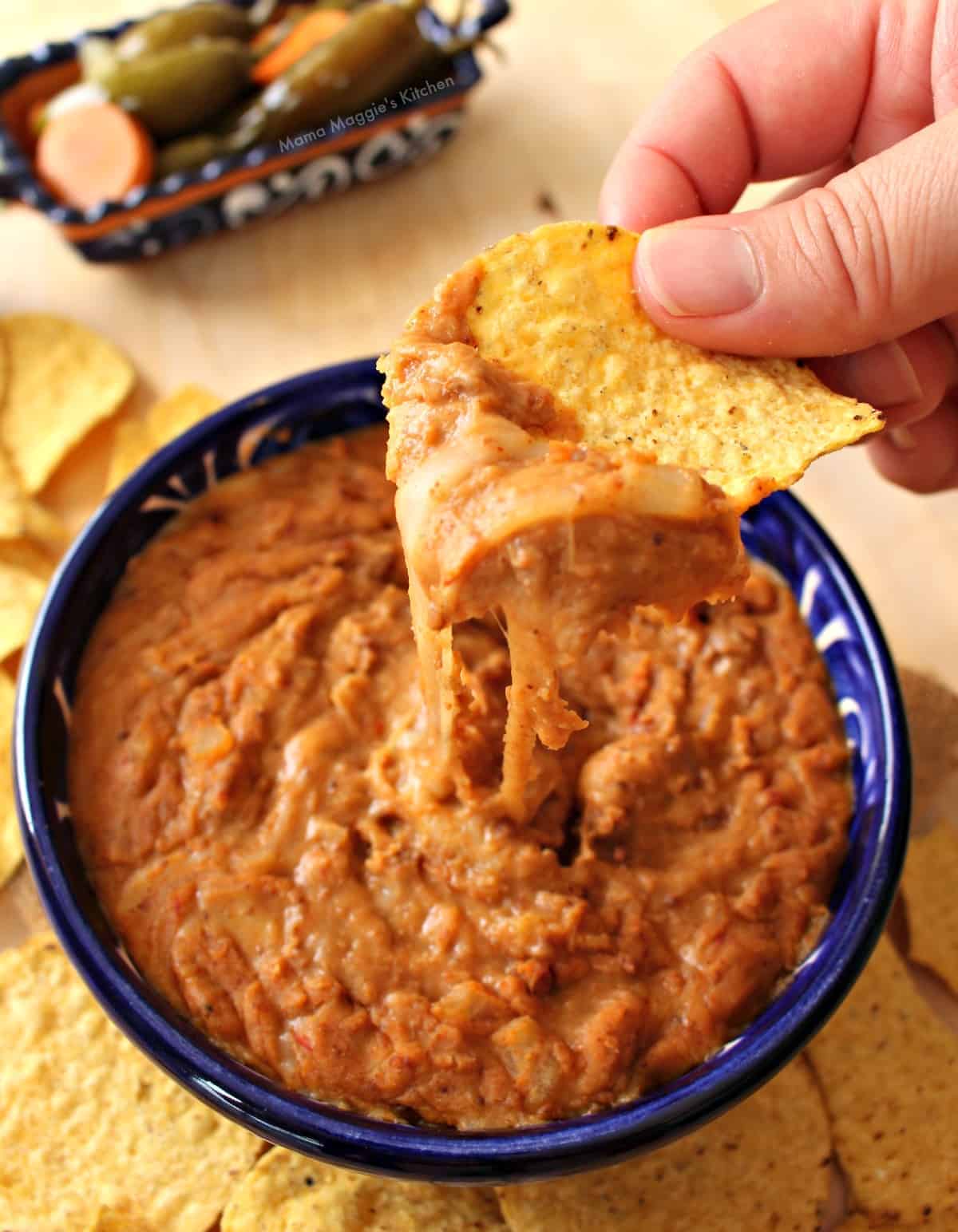 A hand holding a chip with frijoles puercos over a blue bowl.