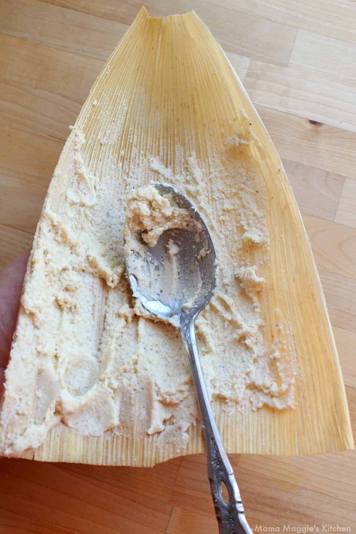 A spoon spreading masa on a corn husk.