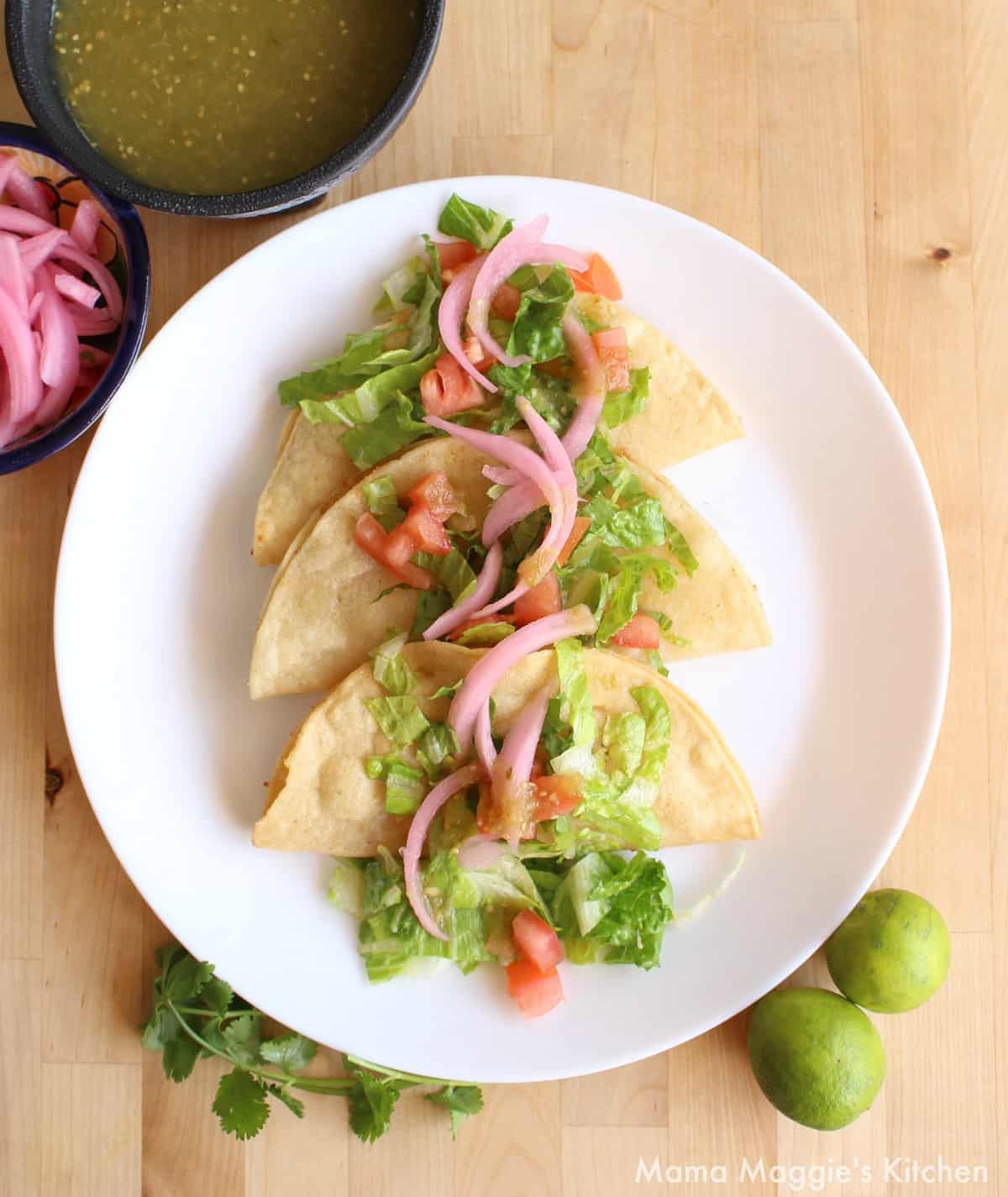 Potato Tacos Dorados on a white plate topped with green lettuce, tomatoes, and pickled red onions.