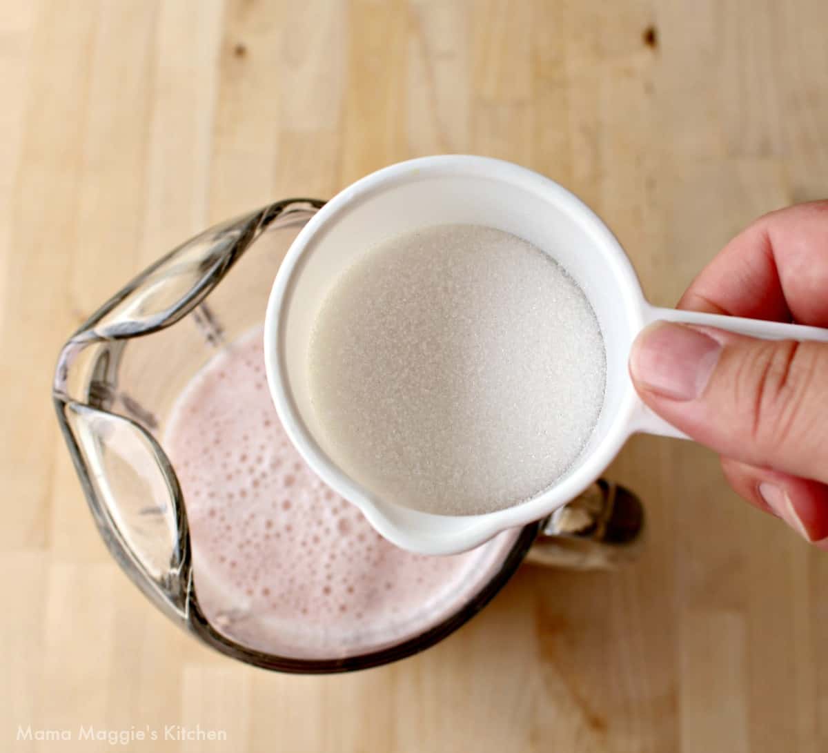 A hand holding a white measuring cup filled with sugar over a large pitcher.