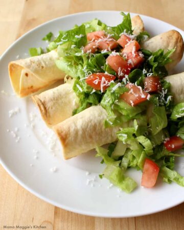 Three flautas topped with lettuce and tomatoes on a white plate.