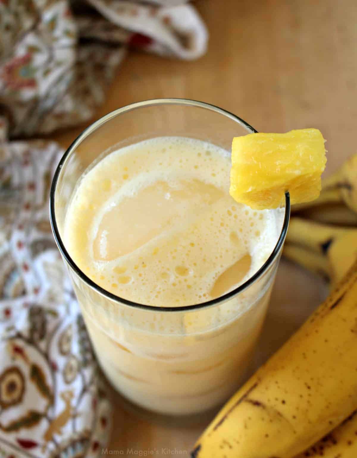 A glass of pineapple banana agua fresca decorated with a pineapple wedge and surrounded by bananas.