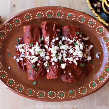 Enchiladas Rojas on a decorative Mexican plate topped with cilantro, crumbled cheese, and diced onion.