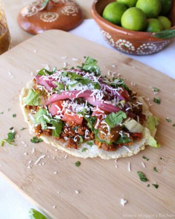 A huarache on a wooden cutting board and topped with meat, lettuce, tomatoes, and cheese.