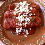 Enchiladas Rojas on a decorative Mexican plate topped with cilantro, crumbled cheese, and diced onion.