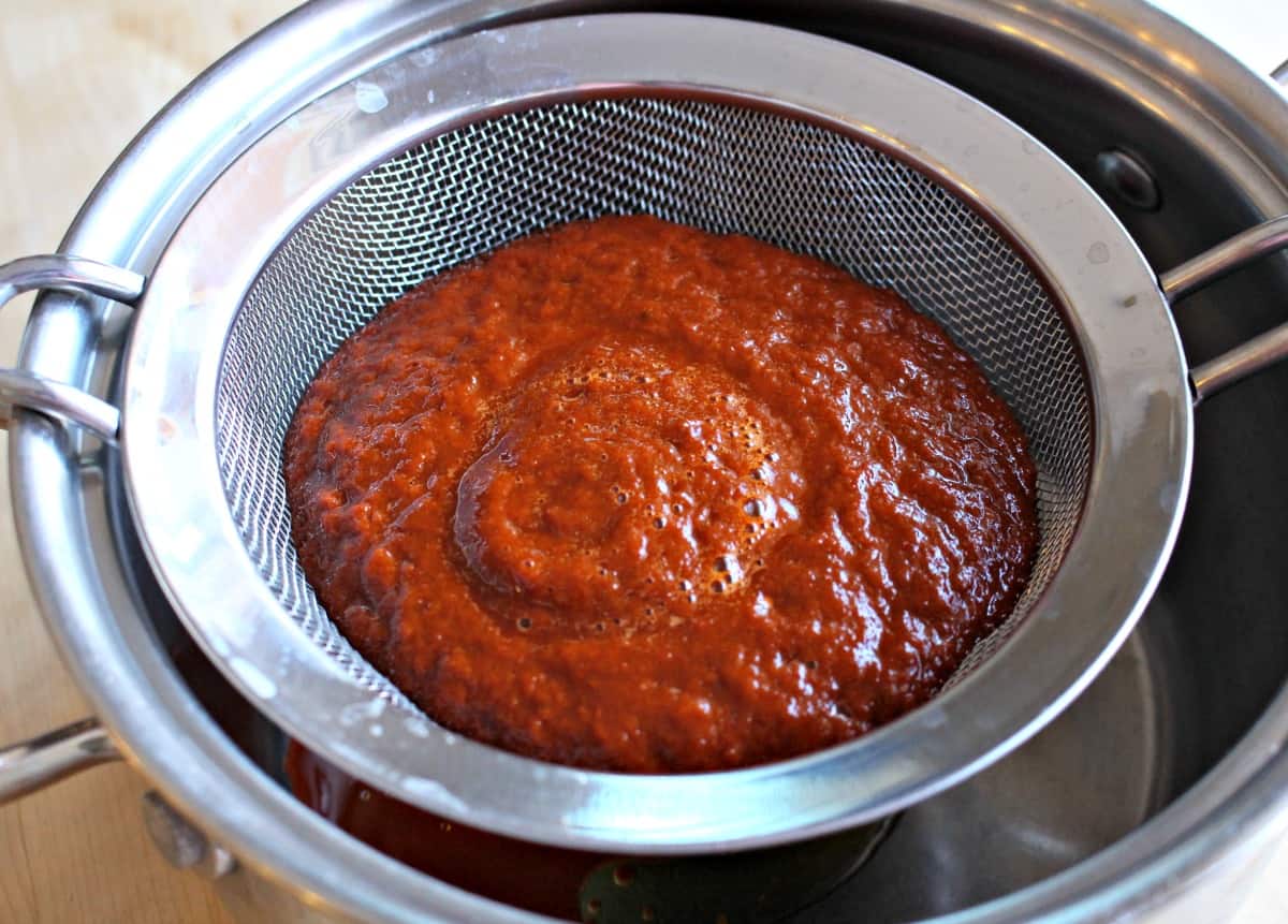 Red sauce strained through a strainer over a metal stock pot.