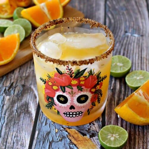A skinny orange margarita in a glass decorated with a sugar skull and surrounded by lime and orange wedges.