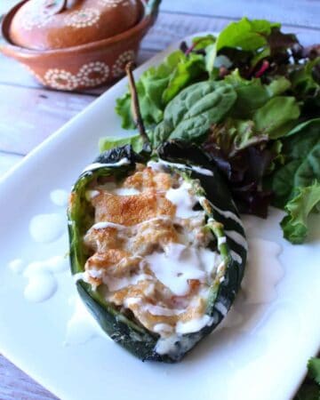 A pork chile relleno on a white plate drizzled with crema mexicana and next to a bed of lettuce.