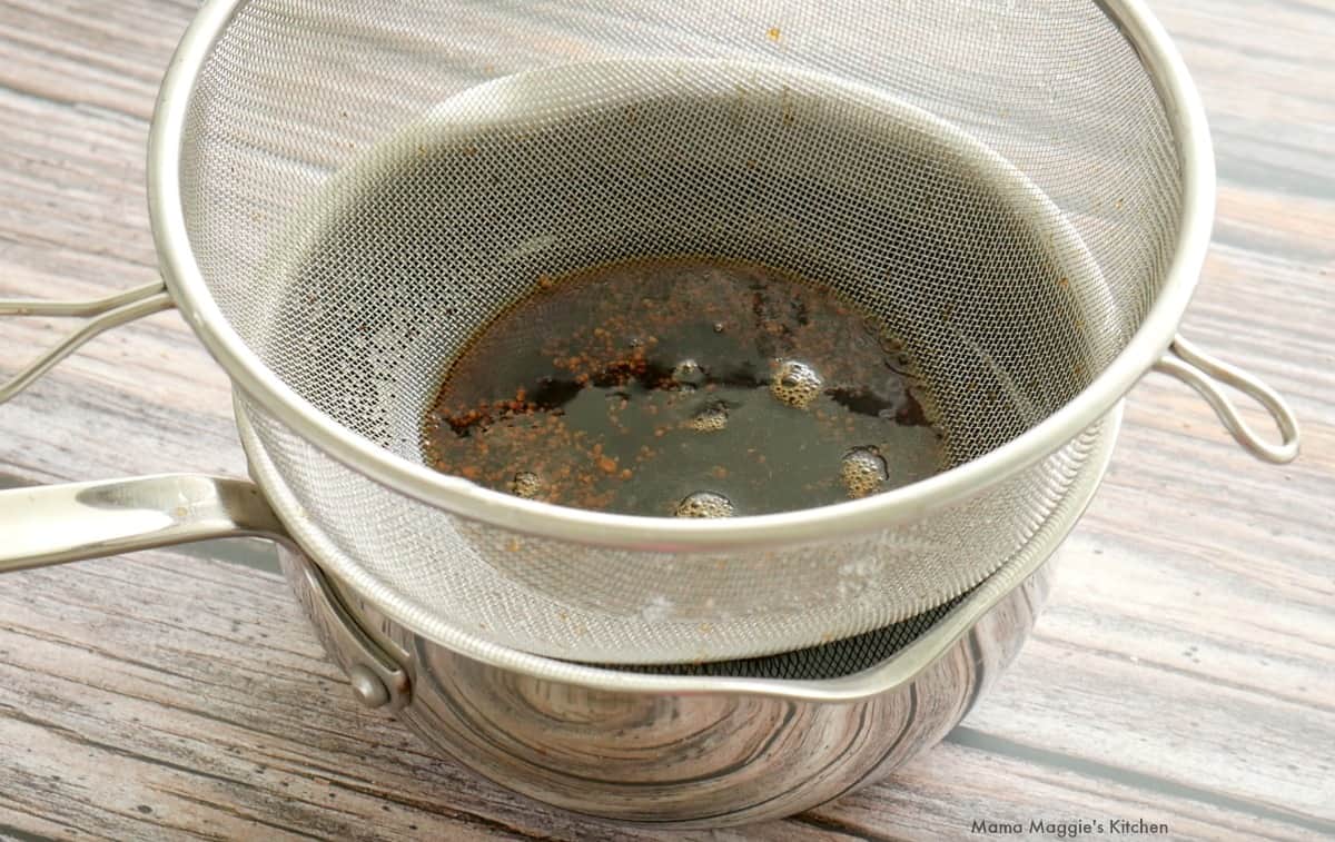 A strainer over a pot removing the cinnamon stick and ground coffee.