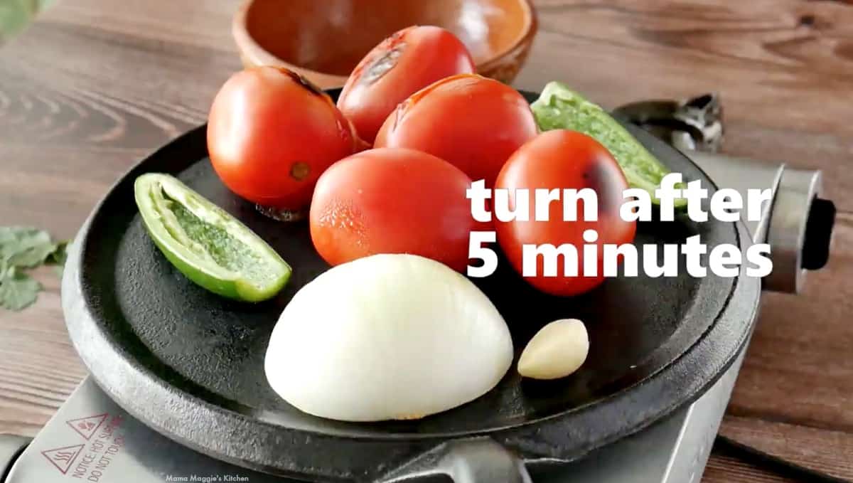 Tomatoes, jalapeno, onion, and garlic roasting on a black skillet.