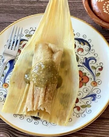 A chicken tamal on a corn husk and on a plate next to a fork and salsa.