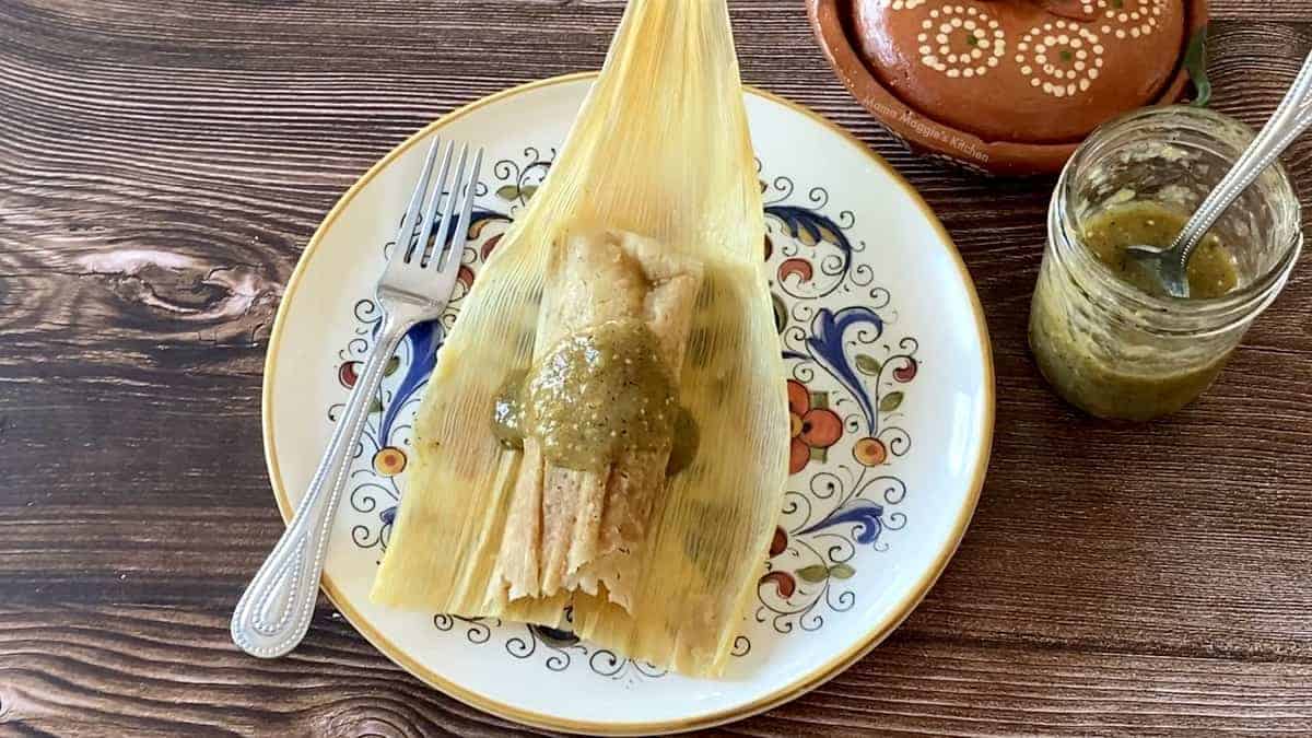 A chicken tamal on a corn husk and on a plate next to a fork and salsa.