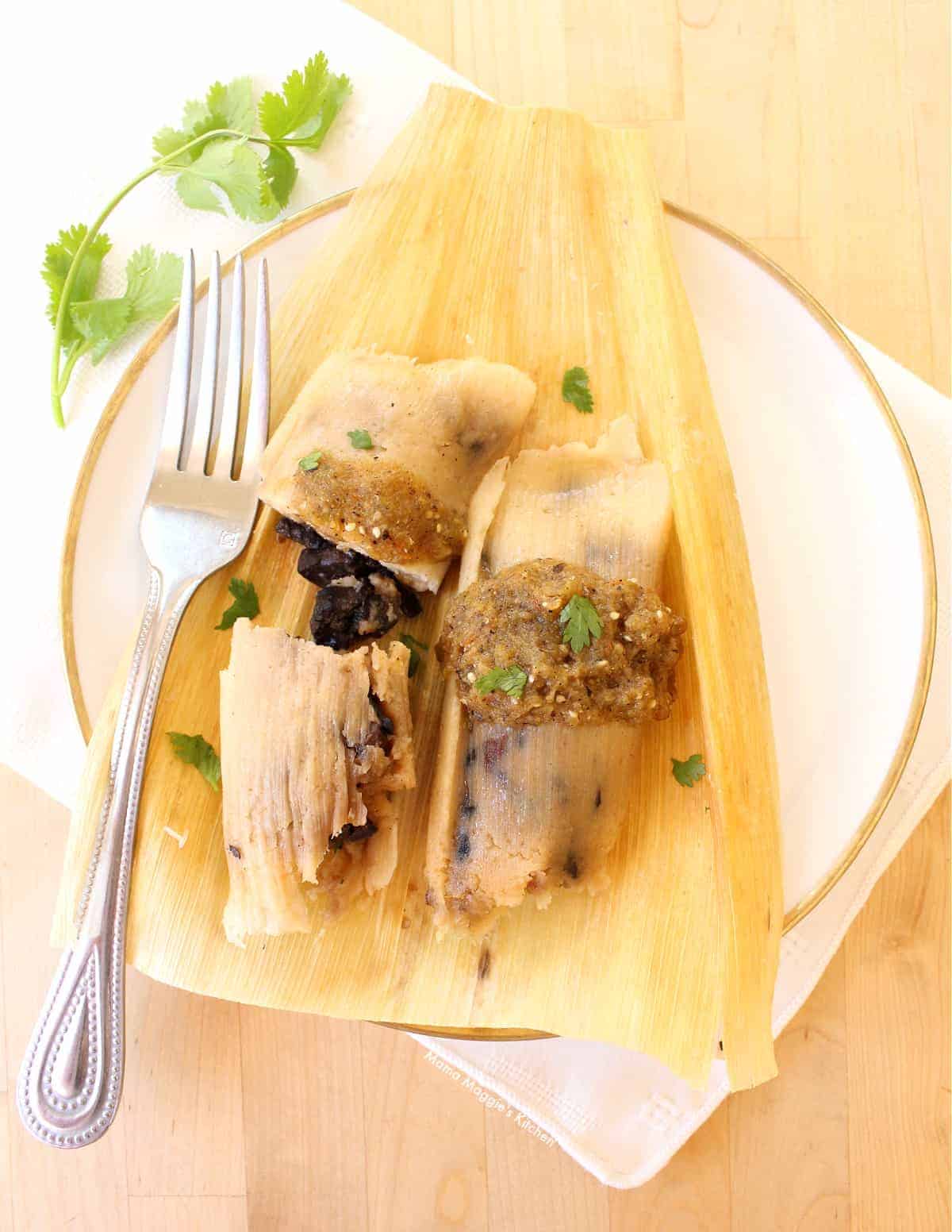Two tamales on a corn husk topped with salsa, cilantro, and served with a fork.