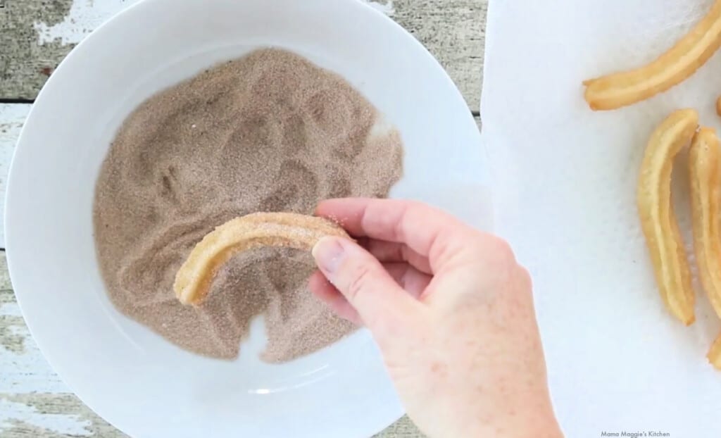 A hand holding a churros over a bowl with cinnamon and sugar.