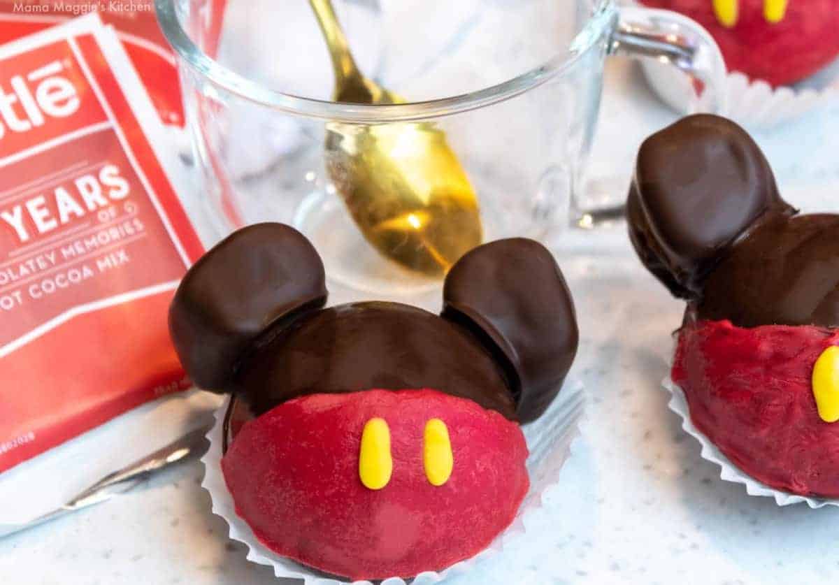 Two Mickey Mouse Hot Chocolate Bombs next to each other next to cocoa mix and a cup.