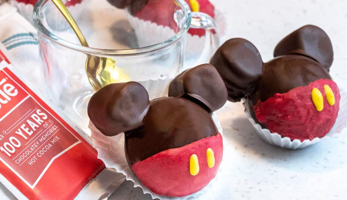 Two Mickey Mouse Hot Chocolate Bombs next to each other next to a glass cup.