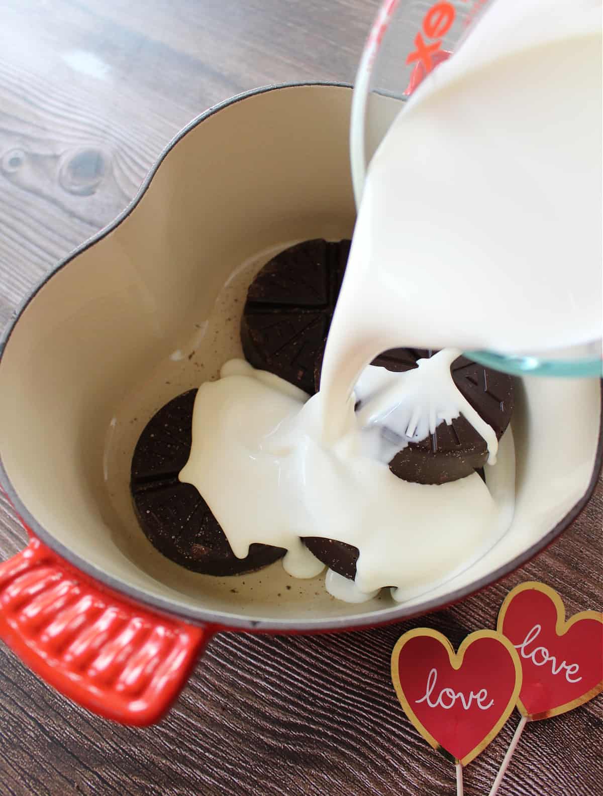 A measuring cup pouring heavy cream into a pot with chocolate.