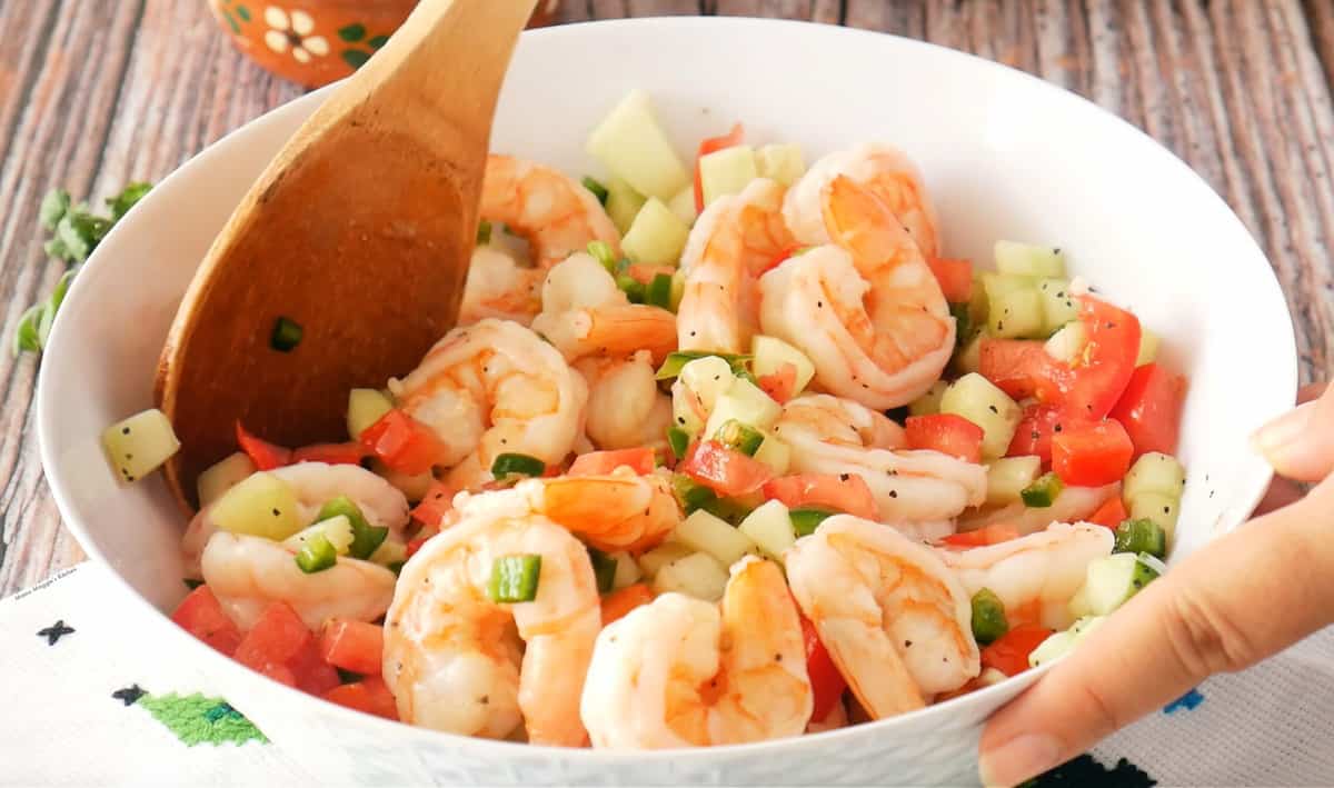 A hand holding a white bowl and mixing the shrimp with a wooden spoon with the other hand.