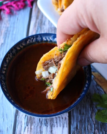 A hand dipping a birria taco into the consomme.