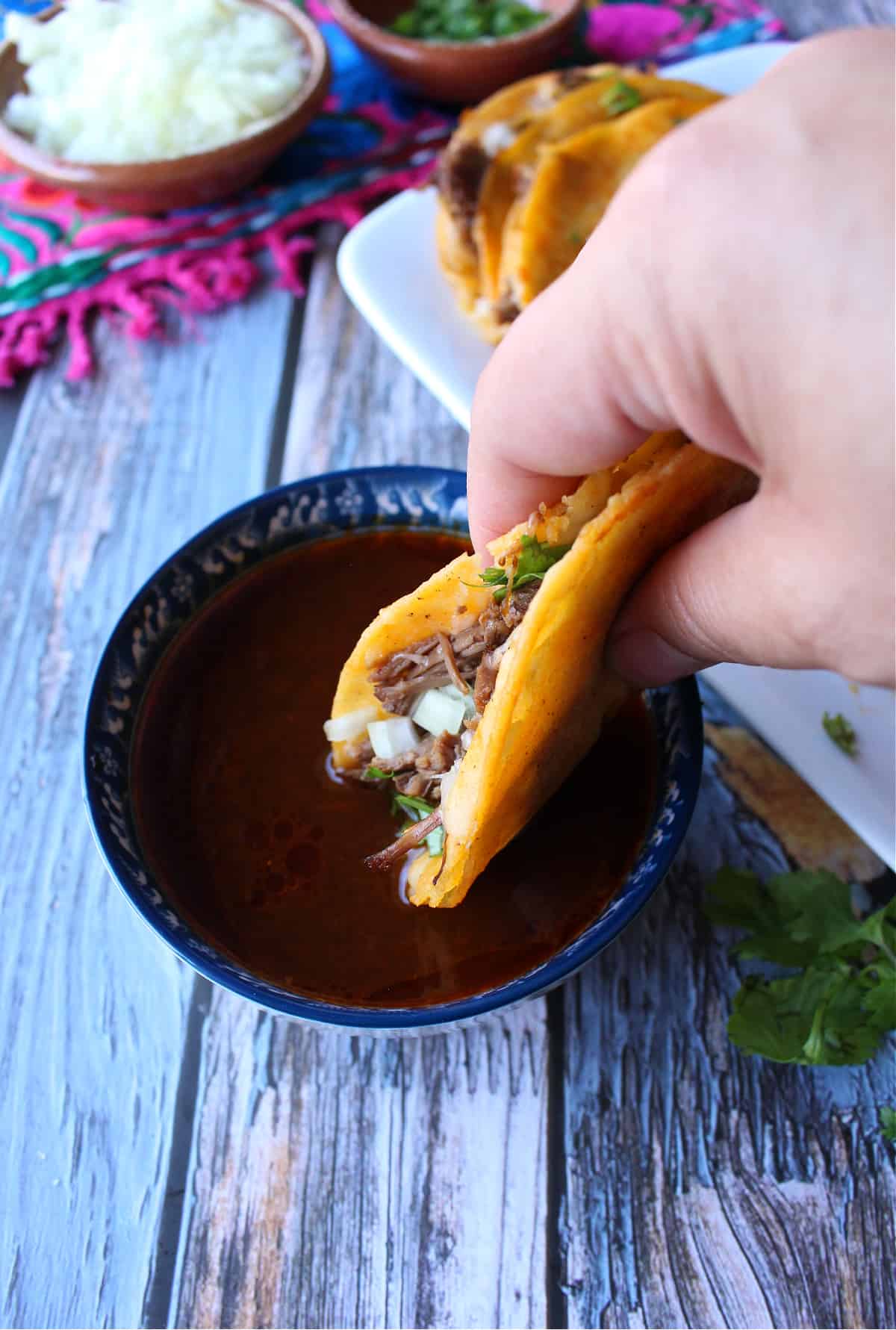A hand dipping a birria taco into the consomme.
