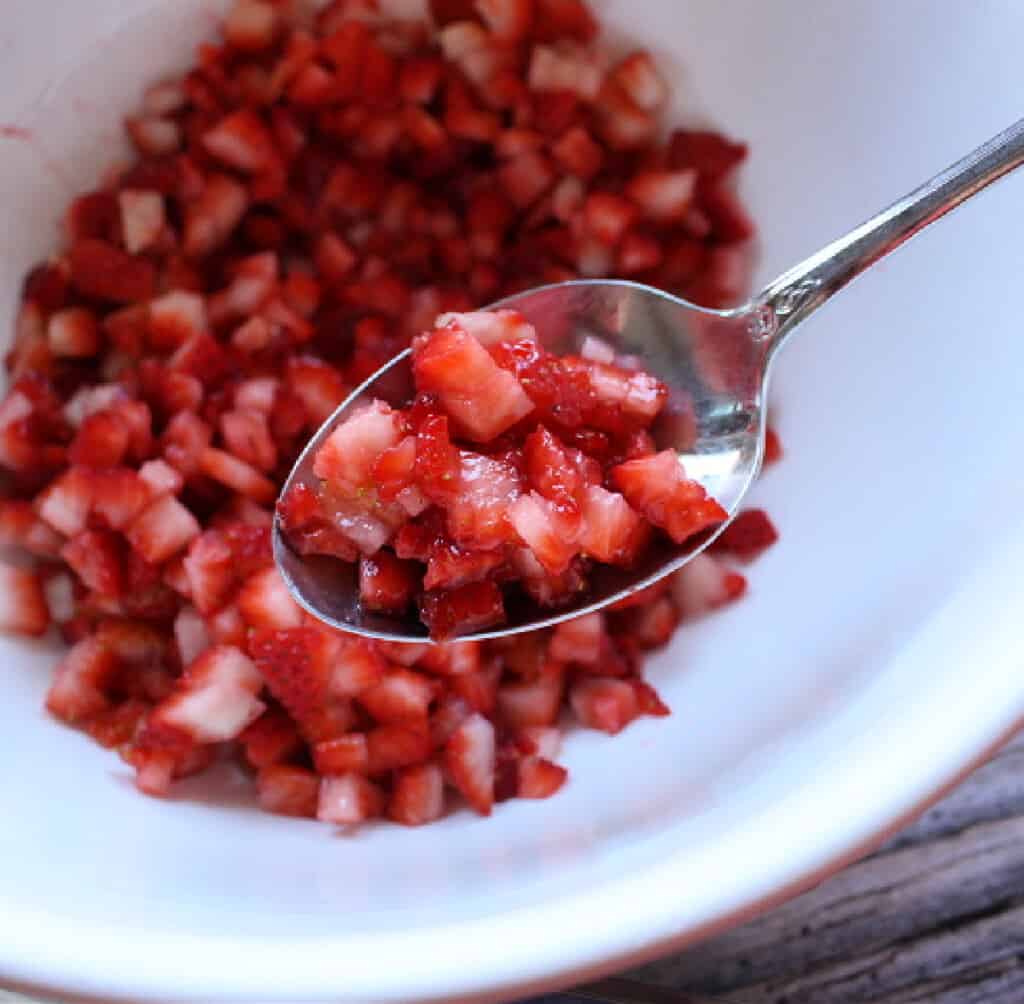 A spoon holding chopped up strawberries over a bowl with more chopped strawberries.