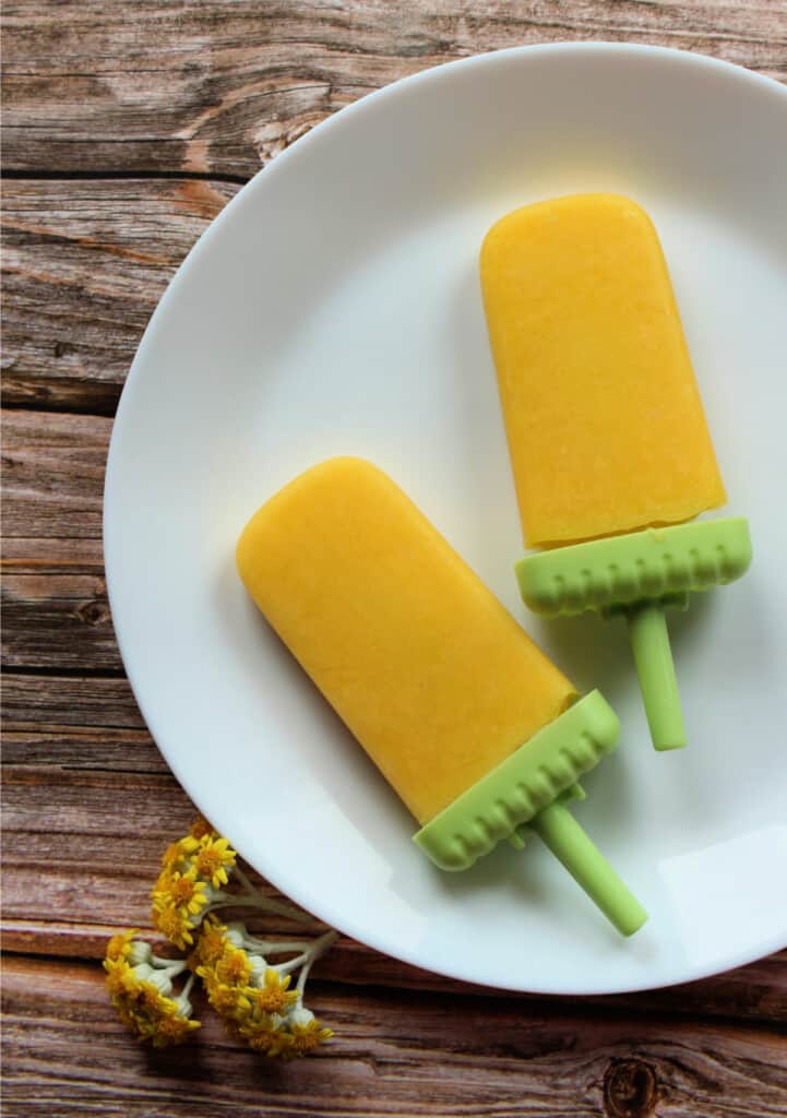 Two Mexican mango paletas sitting on a white plate next to yellow flowers.