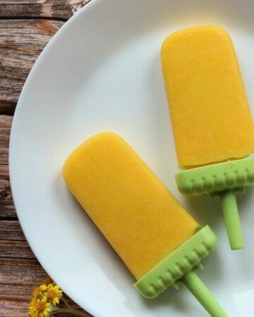 Two Mexican mango paletas sitting on a white plate next to yellow flowers.