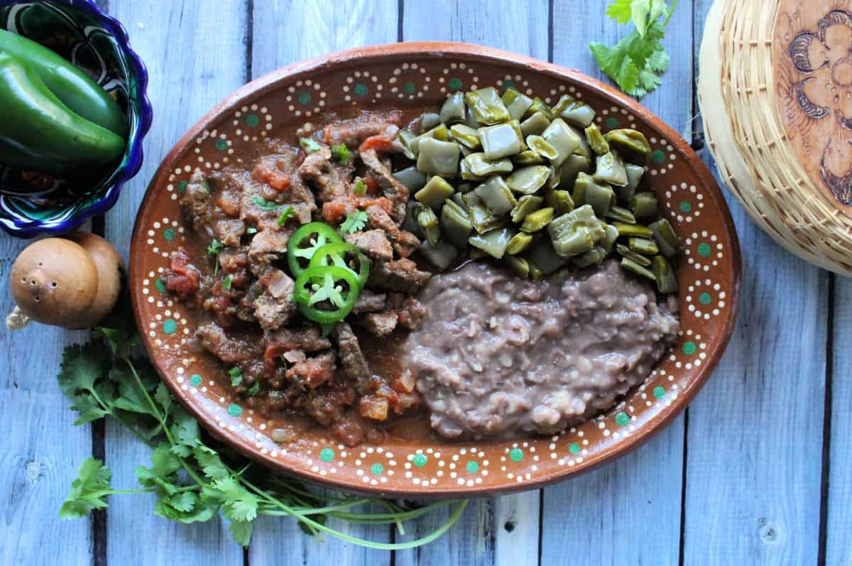 Bistec a la Mexicana served on a decorative clay plate next to beans and nopales.