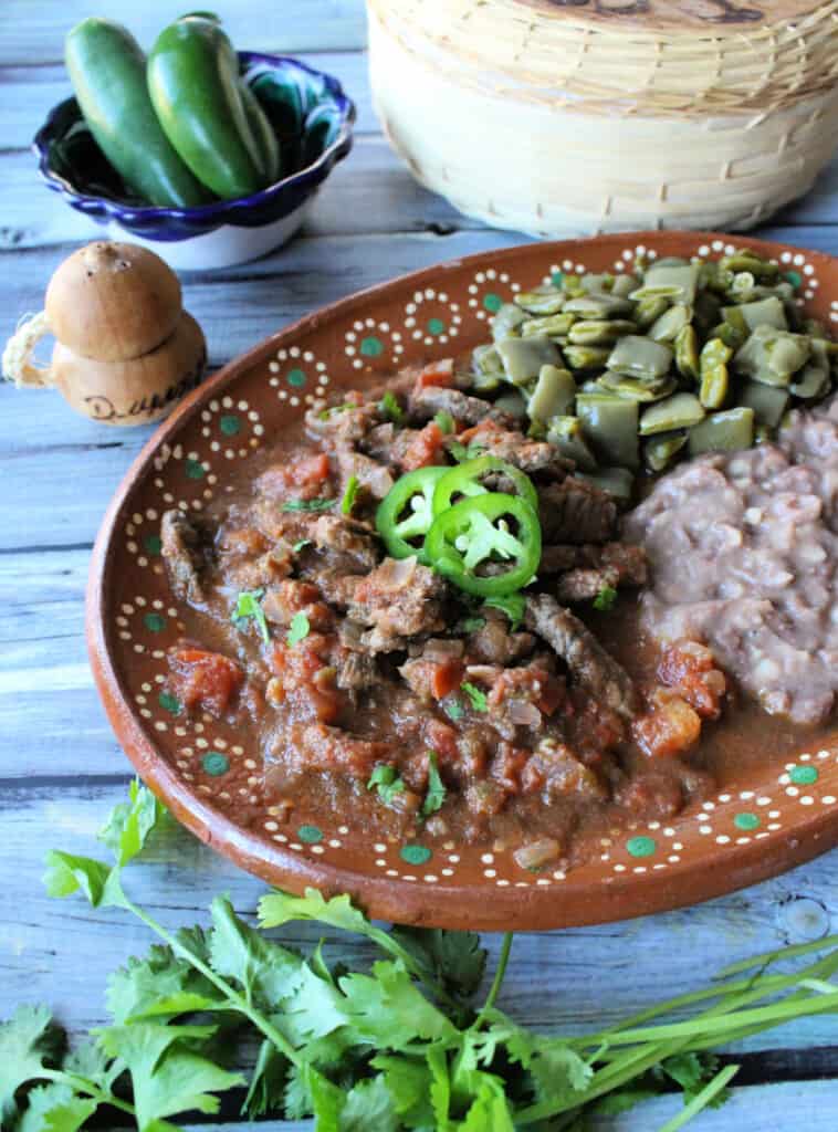 Bistec a la Mexicana served on a decorative clay plate next to beans and nopales.