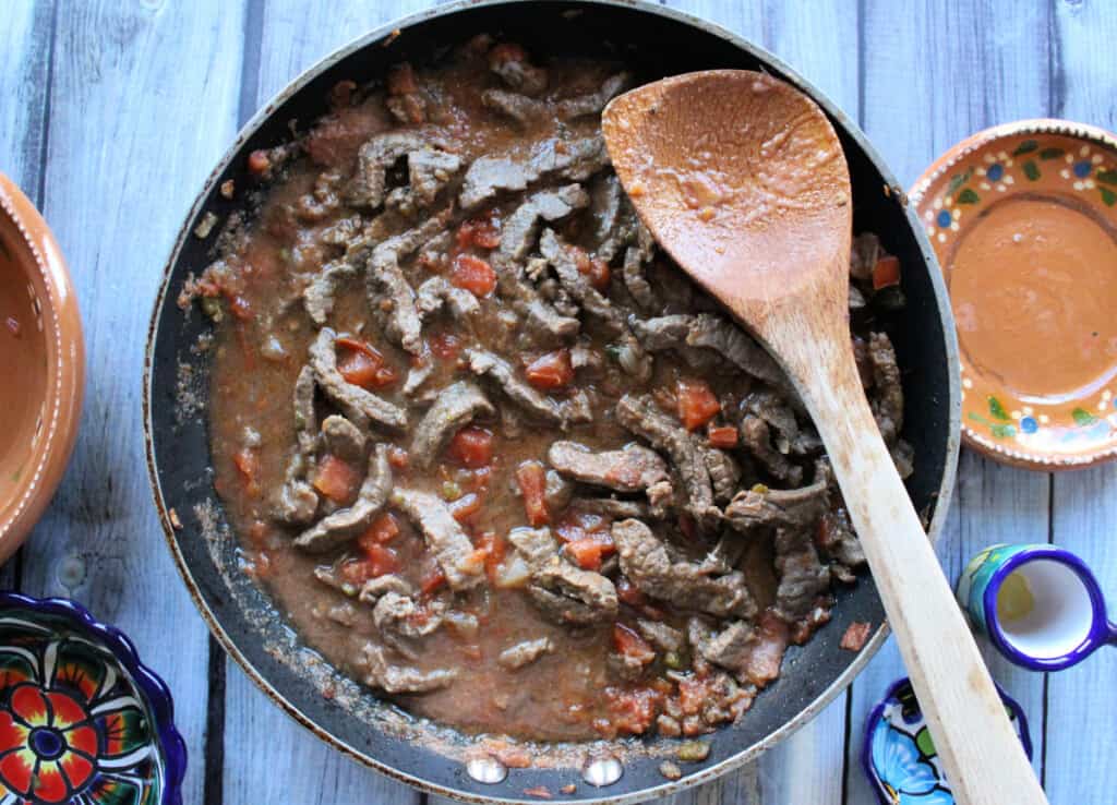 Cooked beef strips in a skillet with a wooden spoon.