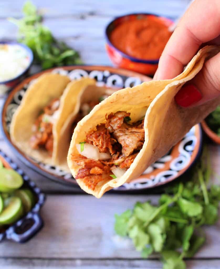 A hand holding a pork taco over a plate with more tacos.