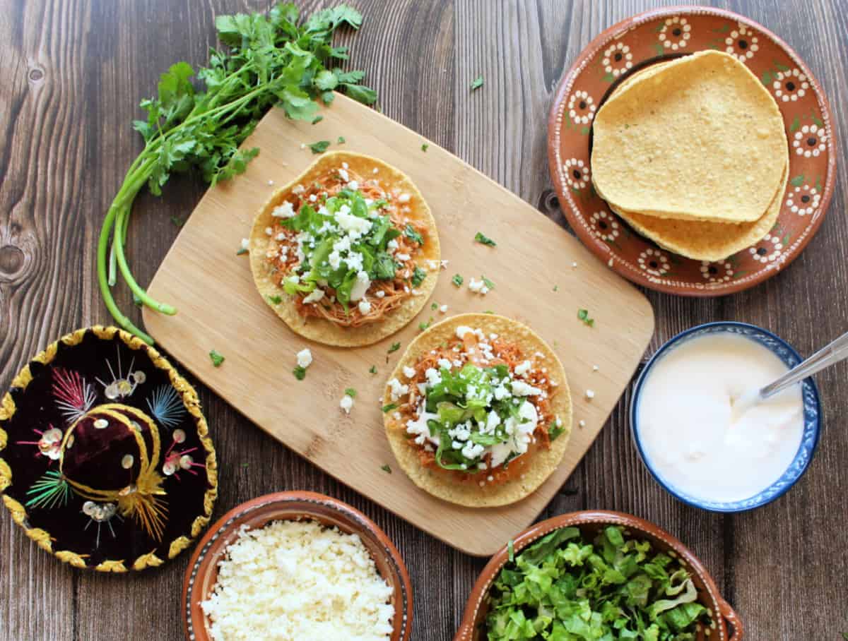 Two tostadas of Tinga de Pollo (Chicken Tinga) on a wooden board next to the toppings.